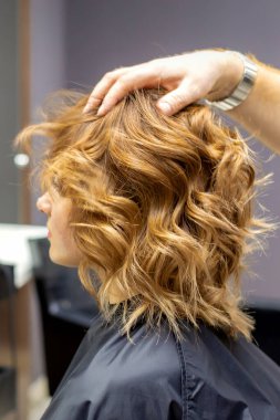 Erkek kuaförün yan görünüşü, kuafördeki genç beyaz kadının kıvırcık saç stilini kontrol ediyor.