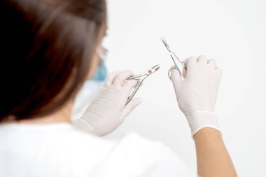 Beyaz maske takan ve beyaz arka planda eldiven takan kadın manikürcünün elindeki manikür aletleri.