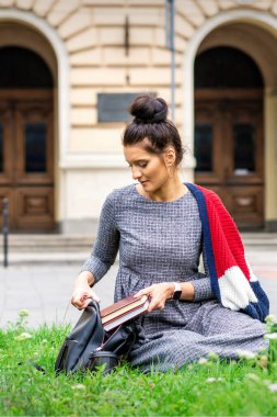 Genç yetişkin kız öğrenci, üniversite binasının yakınındaki çimlerin üzerinde oturan kitapları sırt çantasına koyar.