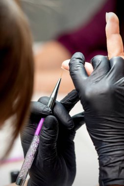 Manikür ustası, manikür salonundaki kadın tırnağının kenarına mor jel kalemle süslemeler yapıyor.