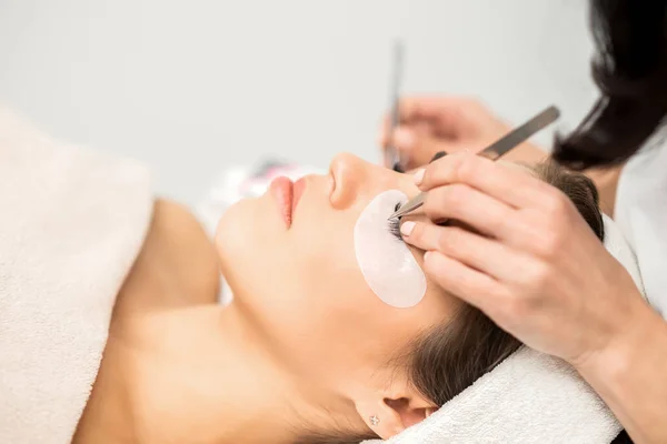 Young caucasian woman having eyelash extension procedure in beauty salon. Beautician glues eyelashes with tweezers