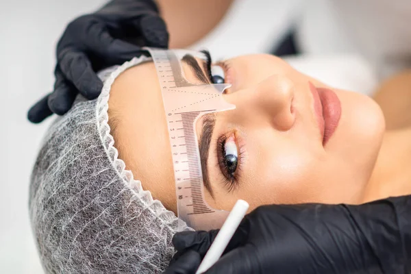 Cosmetologist is measuring with ruler the brows of young caucasian woman before permanent makeup tattoo