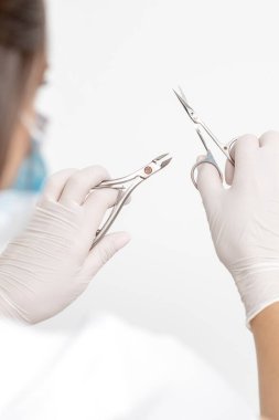 Beyaz maske takan ve beyaz arka planda eldiven takan kadın manikürcünün elindeki manikür aletleri.