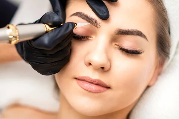 Beautician applying permanent makeup on eyebrows of young woman by special tattoo machine tool