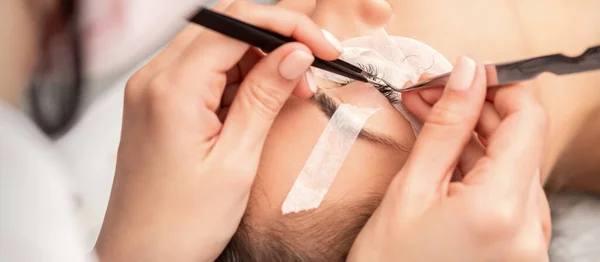 Young caucasian woman having eyelash extension procedure in beauty salon. Beautician glues eyelashes with tweezers