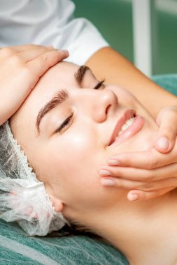 Güzellik salonunda güzellik uzmanları tarafından yüz masajı yaptıran genç bir kadın.