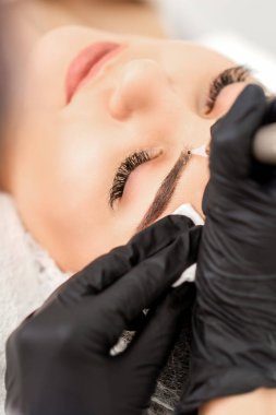 Genç bir kadının kaşlarına özel dövme makinesiyle kalıcı makyaj yapan bir güzellik uzmanı.
