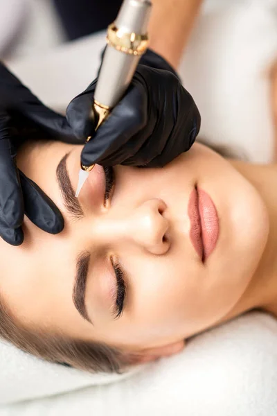 Beautician applying permanent makeup on eyebrows of young woman by special tattoo machine tool