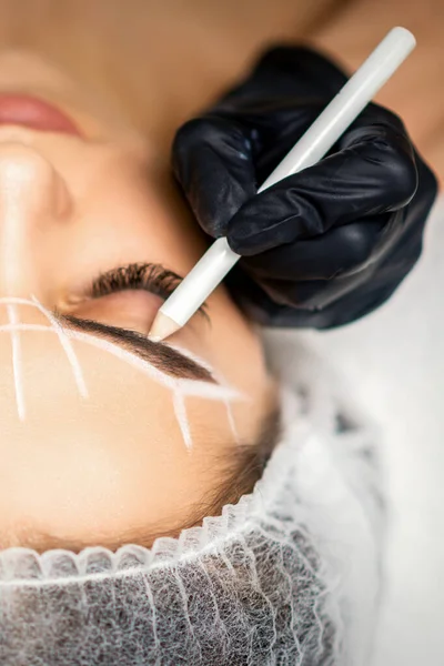 The beautician marks the eyebrow with a white pencil to prepare a permanent makeup procedure