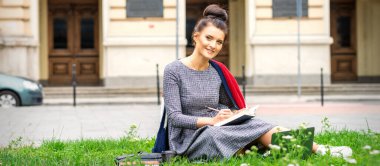 Üniversite binasının yakınındaki çimlerin üzerinde ders çalışan genç ve güzel bir öğrencinin portresi.