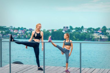 Kadın ve çocuk spor egzersizleri yapıyorlar. Rıhtımda bacaklarını açıyorlar. Gölde.