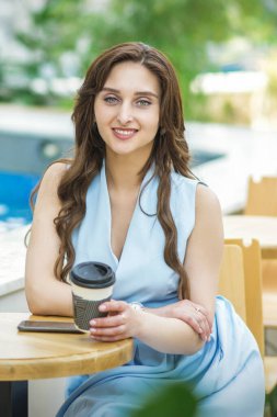 Güzel, genç, beyaz bir kadın havuz kenarında bir fincan sıcak içecekle masada oturuyor.