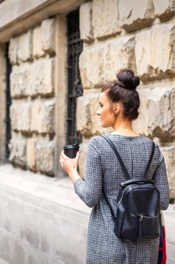 Sıcak bir içecek ve sırt çantasıyla bir Avrupa şehrinde yürüyen genç bir kadının arkası.