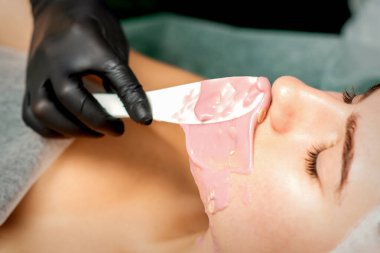 Kozmetik uzmanı güzellik salonundaki genç bir kadının yüzüne aljinik bir maske takıyor.