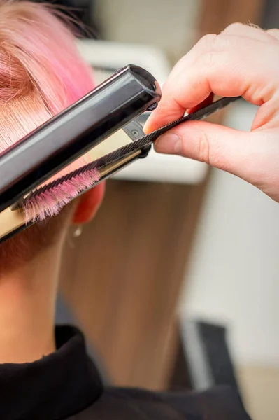 在美容院里 理发师用理发熨斗把女顾客的短粉色头发理直 — 图库照片
