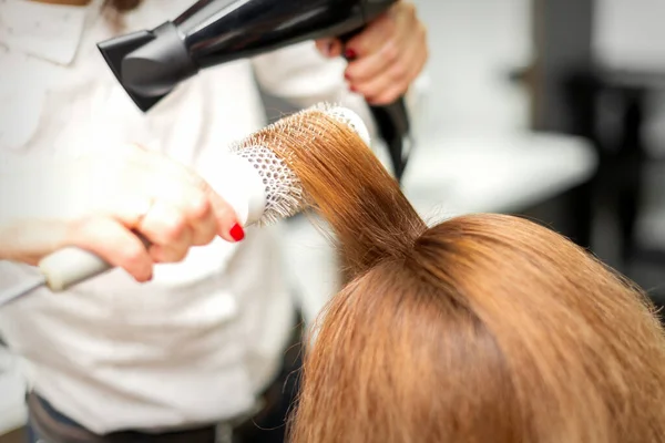 在美容院里用吹风机和圆形刷子擦干长长的红头发的理发师的近照 — 图库照片