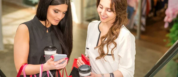 两个漂亮的高加索年轻女人在购物中心里看着装有手提包的智能手机 — 图库照片