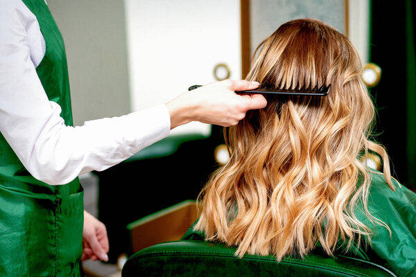 Back view of hairdresser combing wavy hair of a young blonde woman in a beauty salon