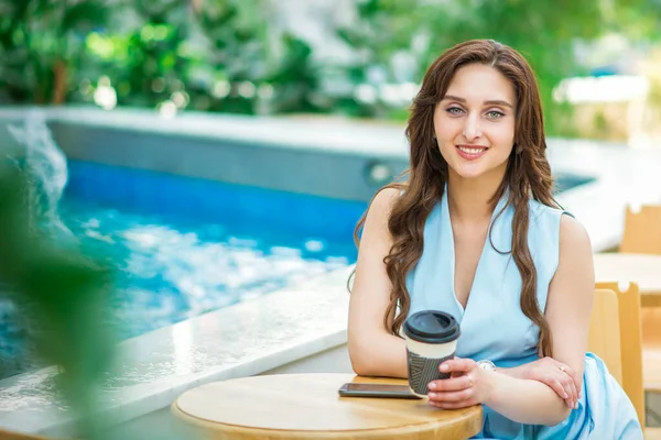 プールの近くに熱い飲み物のカップでテーブルに座っている美しい若い白人女性 — ストック写真