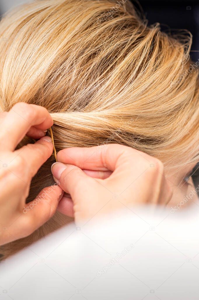 Closeup back view of a hairdresser stabs blonde hair with a hairpin for a young woman in a hair salon