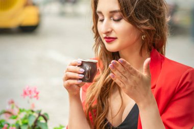 Güzel genç bir kadın küçük bir fincanda kahve kokluyor ve açık bir kafedeki masada oturup kahve içiyor.