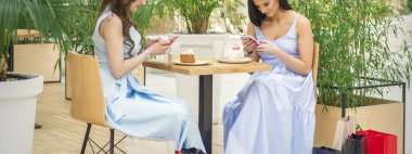 Two young caucasian women sitting at the table and looking at the smartphones with shopping bags in a cafe outdoors