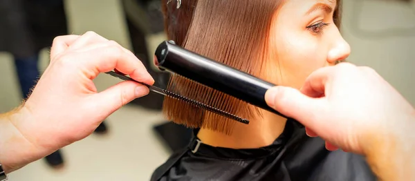 Hair stylist\'s hands straightening short hair of young brunette woman with flat iron and comb in a beauty salon