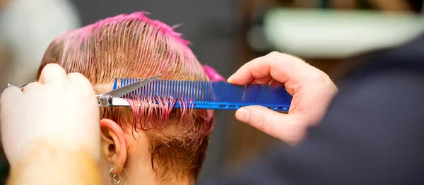 一头粉色头发的年轻的高加索女人在理发店里被一个男理发师的手剪短了头发 — 图库照片