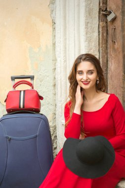 Beyaz bir kadın, elinde bavuluyla eski bir evin kapısında oturuyor. Dışarıdaki kameraya bakıyor.