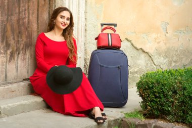 Beyaz bir kadın, elinde bavuluyla eski bir evin kapısında oturuyor. Dışarıdaki kameraya bakıyor.