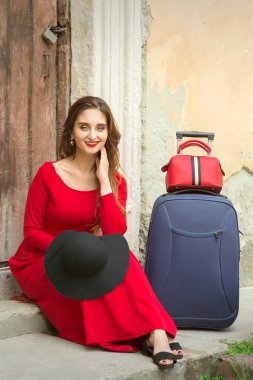 Beyaz bir kadın, elinde bavuluyla eski bir evin kapısında oturuyor. Dışarıdaki kameraya bakıyor.