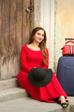 Beyaz bir kadın, elinde bavuluyla eski bir evin kapısında oturuyor. Dışarıdaki kameraya bakıyor.