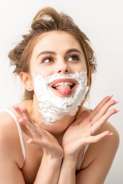 Yüzünde tıraş köpüğü olan güzel, genç, gülümseyen beyaz bir kadının portresi beyaz arka planda dilini dışarı çıkardı.