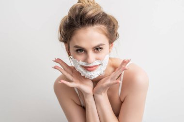 Yüzünde tıraş köpüğü olan güzel, gülümseyen beyaz bir kadının portresi beyaz arka planda poz veriyor.