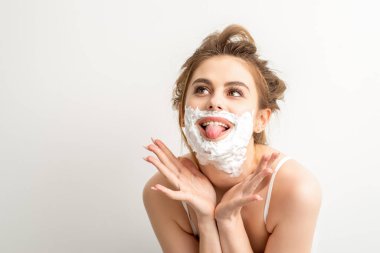 Yüzünde tıraş köpüğü olan güzel, genç, gülümseyen beyaz bir kadının portresi beyaz arka planda dilini dışarı çıkardı.