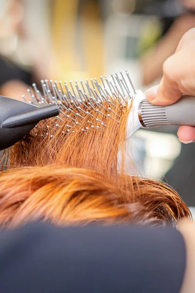 Secagem de cabelo no estúdio de cabeleireiro. estilista de cabeleireira  seca o cabelo com secador de cabelo e escova redonda ruiva de uma mulher em  um salão de beleza.