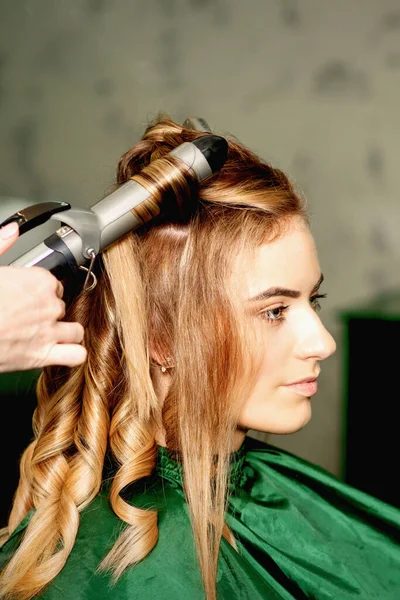 Hairdresser using curling tongs curls long brown hair on the young caucasian girl in a beauty salon