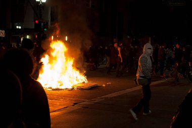 Oakland 'da 9 Kasım 2016' da Donald Trump 'ın seçilmesine karşı yapılan protestolar sırasında Broadway' de çıkan sokak yangınının önünde maskeli bir protestocu duruyor..