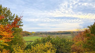 Almanya 'nın kuzeyinde Schleswig-Holstein eyâletinde, Frruper Berge doğa koruma alanında, dramatik bulutlu bir gökyüzü altında yeşil bir vadi ve renkli bir sonbahar ormanı üzerinde panoramik manzara. Manzaralı kırsal alan, doğa arka planı ve huzurlu kırsal alan.