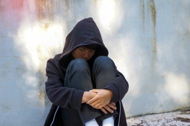 Siyah kapüşonlu üzgün genç bir çocuk kirli bir duvara dizlerini dayayıp depresyon ve stresi sembolize ediyor..                               