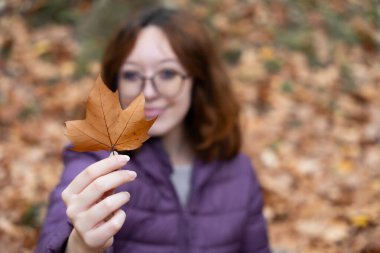 Gülümseyen bir kadın, düşmüş yaprakların bulanık arka planına karşı kahverengi bir sonbahar yaprağı sunuyor.