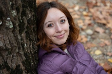 Kızıl saçlı, mor ceketli, açık havada gülümseyen ve ağaca yaslanan genç bir kadın.