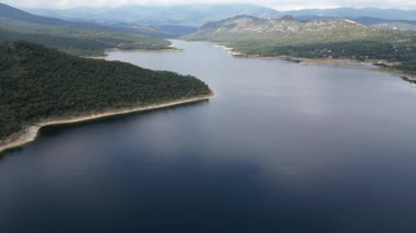 Yüksek açılı drone görüntüleri yeşil bir ormanla çevrili engin bir dağ gölünün üzerinde uçuyor.