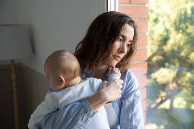 Yeni anne bebeğini kucağında tutuyor ve pencereden dışarı bakıyor. Doğum sonrası depresyonundan bunalmış.