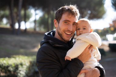 Genç baba oğlunu kucağında tutuyor, güneşli bir günün tadını çıkarıyor. Aile sevgisini ve ebeveynliği temsil ediyor.