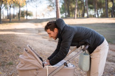 Genç baba gülümsüyor, bebek arabasında bebeğe yaslanıyor. Dışarıda geçirilen sonbahar gününün tadını çıkarmak, aile bağlarını ve ebeveynliği teşvik etmek.