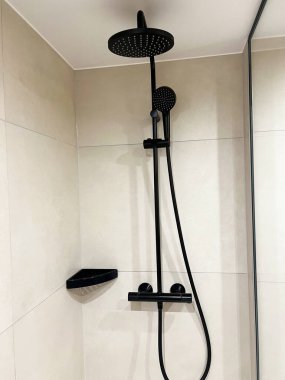 Minimalist duş iç kısmı mat, siyah yağmur duşu ve bej kaplama bir duvarda el yapımı duş başlıkları içeriyor. Köşedeki rafı ve cam bölmesi temiz ve modern bir görünüm için ışığı yansıtan parlak bir tasarım..