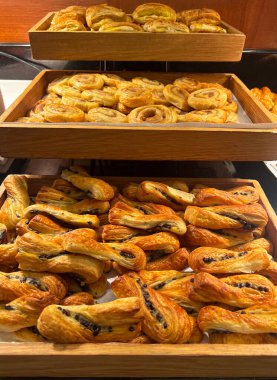 Açık büfedeki ahşap tepsilerde sergilenen taze fırınlanmış hamur işleri. Çeşit çeşit ayçöreği, altın hamurlu kıvrımlar ve girdaplar. Otel ya da kafede fırın kahvaltısı seçimi, lezzetli hamur işleri..