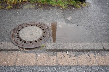 Zarar görmüş caddesi ve kaldırımı olan paslı rögar kapağı