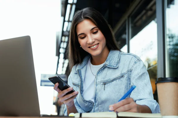 Joven mujer de negocios que trabaja en casa terraza abierta, sentado frente a la computadora portátil, tomando notas en cuaderno negro. — Foto de Stock